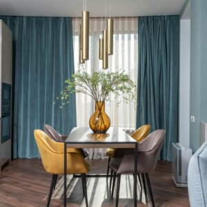 Glass vase with flowers on table with chairs placed near kitchen cabinets and soft couch in modern apartment with window